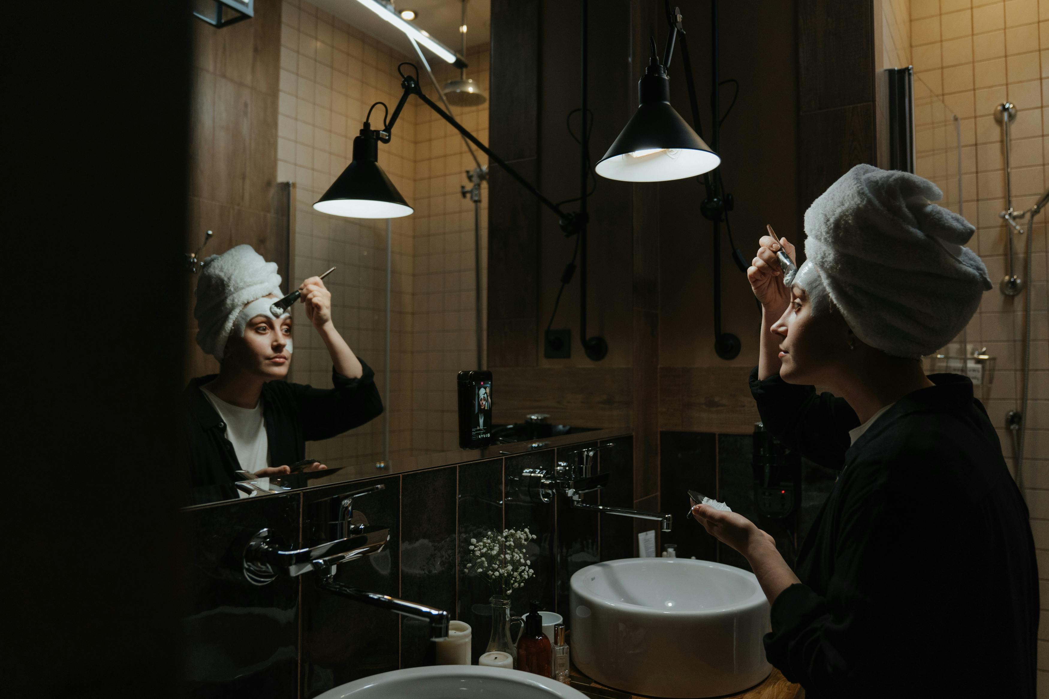 Person with a towel on their head applying makeup in a bathroom.